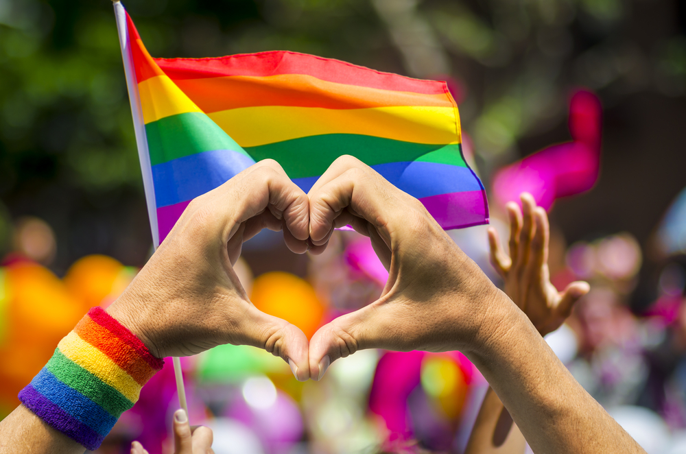 Picture showing the flag of the LGBTQ Community, with two hands that make out a heart.